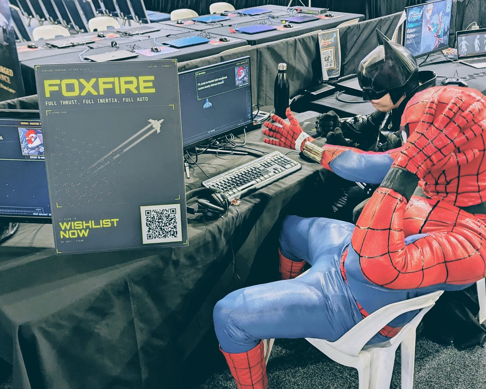 The FOXFIRE booth at Supanova Sydney 2026 &mdash; a Spider-Man cosplayer and a Batman cosplayer play the game at the demo station, beside a FOXFIRE banner reading &lsquo;Full thrust, full inertia, full auto&rsquo; with a wishlist QR code